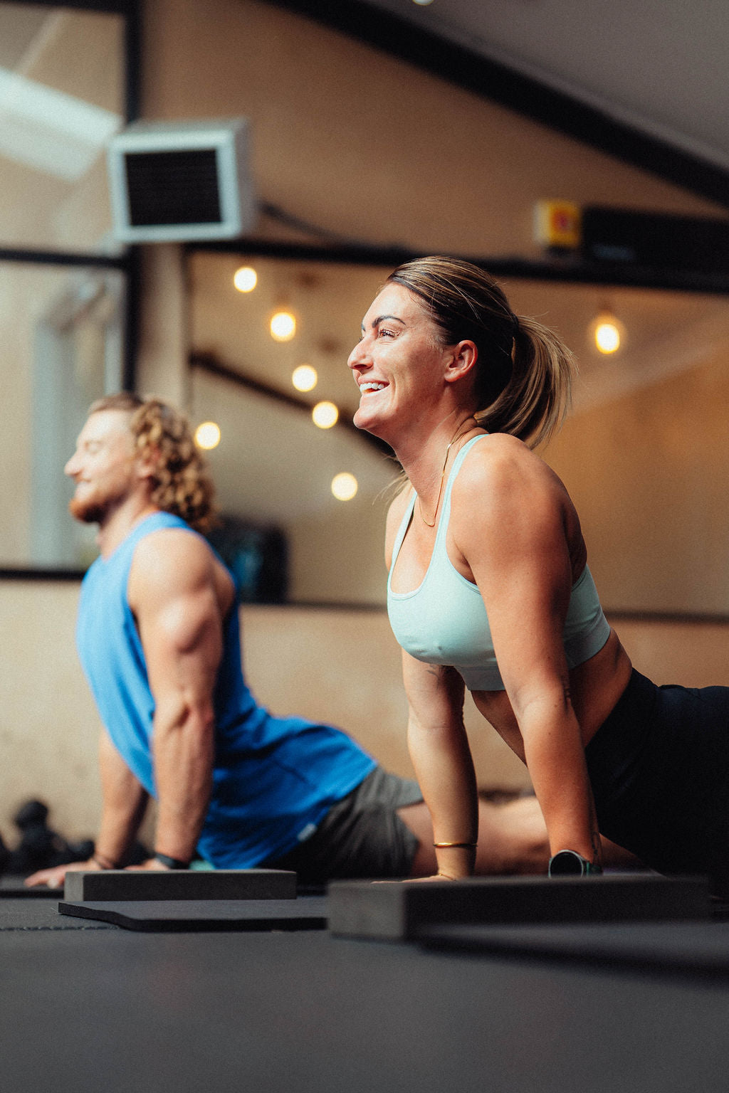 Two people in a gym setting, one in a blue tank top and shorts, the other in a light blue sports bra and black leggings, both doing "Upward-Facing Dog" yoga poses.